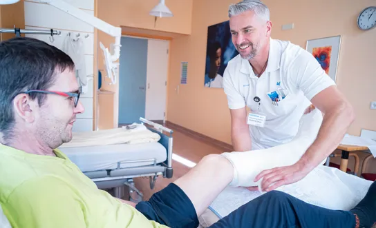 Ein Arzt behandelt in einem Krankenhauszimmer einen im Bett liegenden Patienten, der am linken Unterschenkel und Fuß einen Verband trägt. Ein Arzt behandelt in einem Krankenhauszimmer einen im Bett liegenden Patienten, der am linken Unterschenkel und Fuß einen Verband trägt.